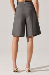 A person is wearing high-waisted gray shorts made of a textured fabric with a tailored fit. The shorts have a waistband with belt loops, a center back seam, and two horizontal slit pockets at the back. The hem falls just above the knees. The person is also wearing beige high-heeled shoes with a pointed toe and slim heel.