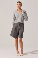 A woman is wearing a light gray, knitted cardigan with pearl buttons, featuring a textured pattern on the shoulders. She is also dressed in high-waisted, gray tailored shorts with pleats and a front zipper, paired with beige pointed-toe heels.