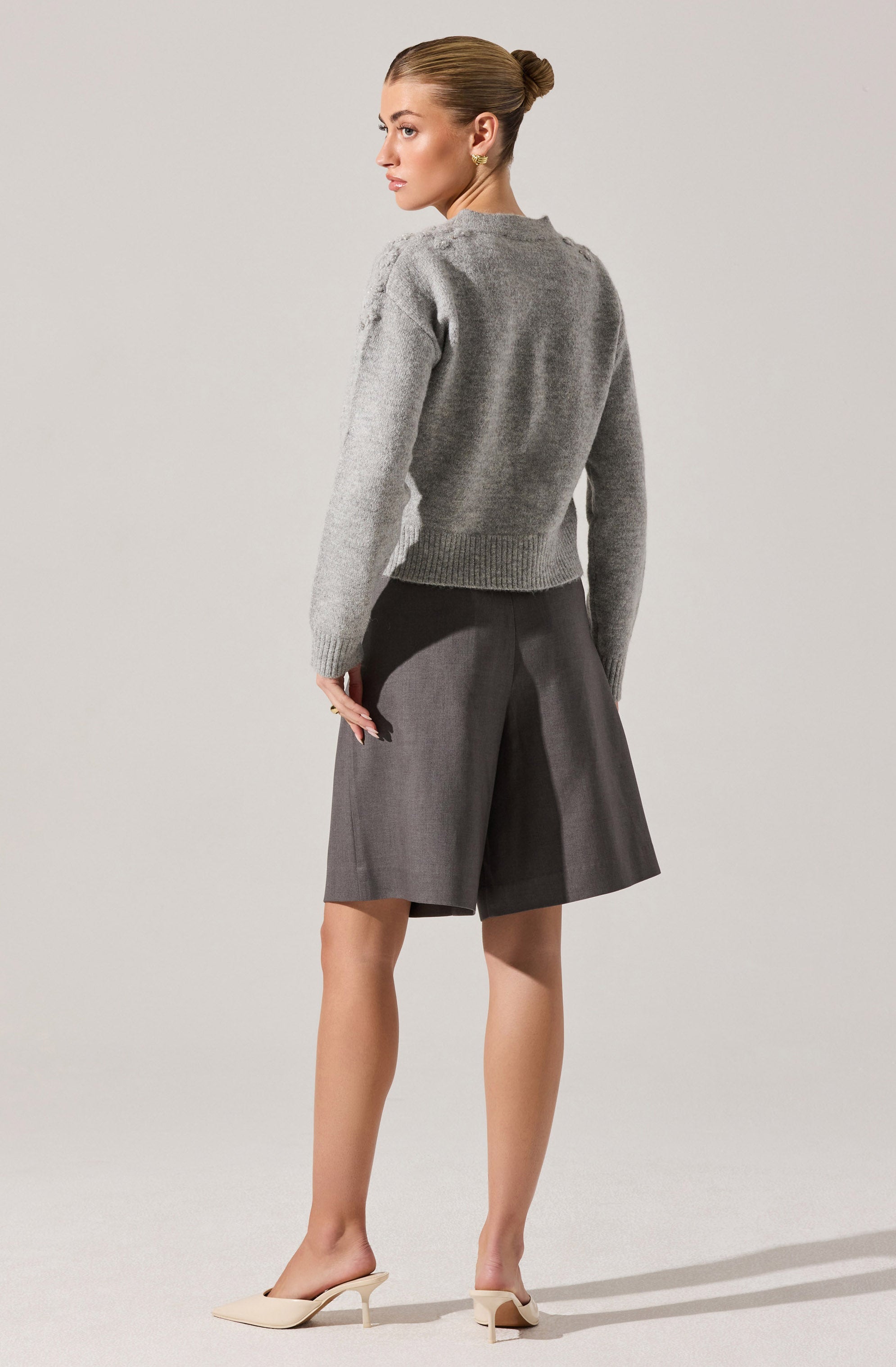 A woman is wearing a light gray, long-sleeved knit sweater with a ribbed waistband. She pairs it with high-waisted, wide-leg dark gray shorts that fall just above the knees. She completes her outfit with a pair of beige, pointed-toe mules with a low, kitten heel.