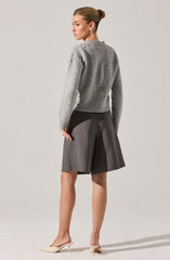 A woman is wearing a light gray, long-sleeved knit sweater with a ribbed waistband. She pairs it with high-waisted, wide-leg dark gray shorts that fall just above the knees. She completes her outfit with a pair of beige, pointed-toe mules with a low, kitten heel.