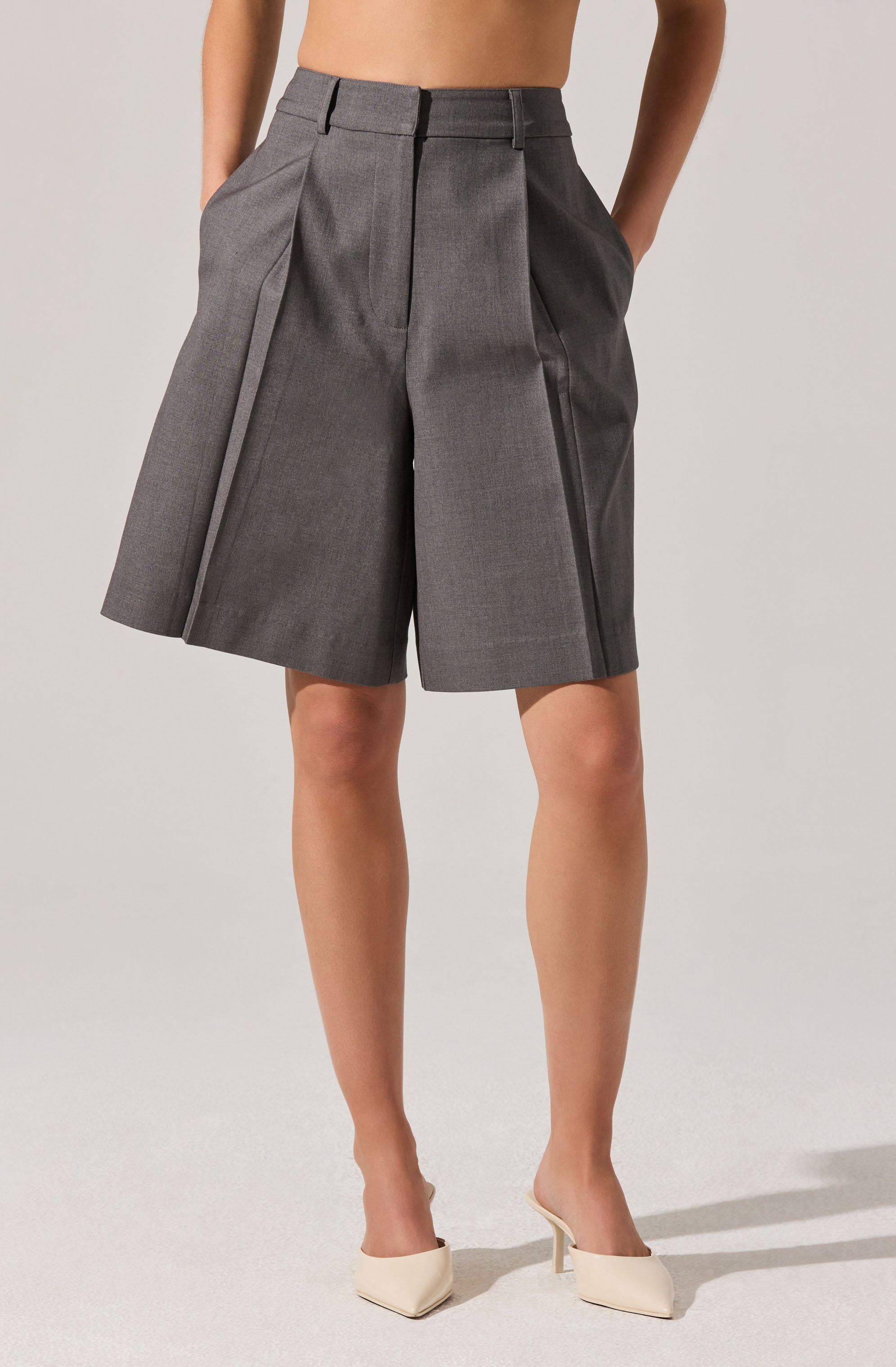 A person is wearing high-waisted, tailored gray shorts with pleats and decorative stitching along the front. The shorts have belt loops and side pockets. They are paired with beige pointed-toe heeled shoes.