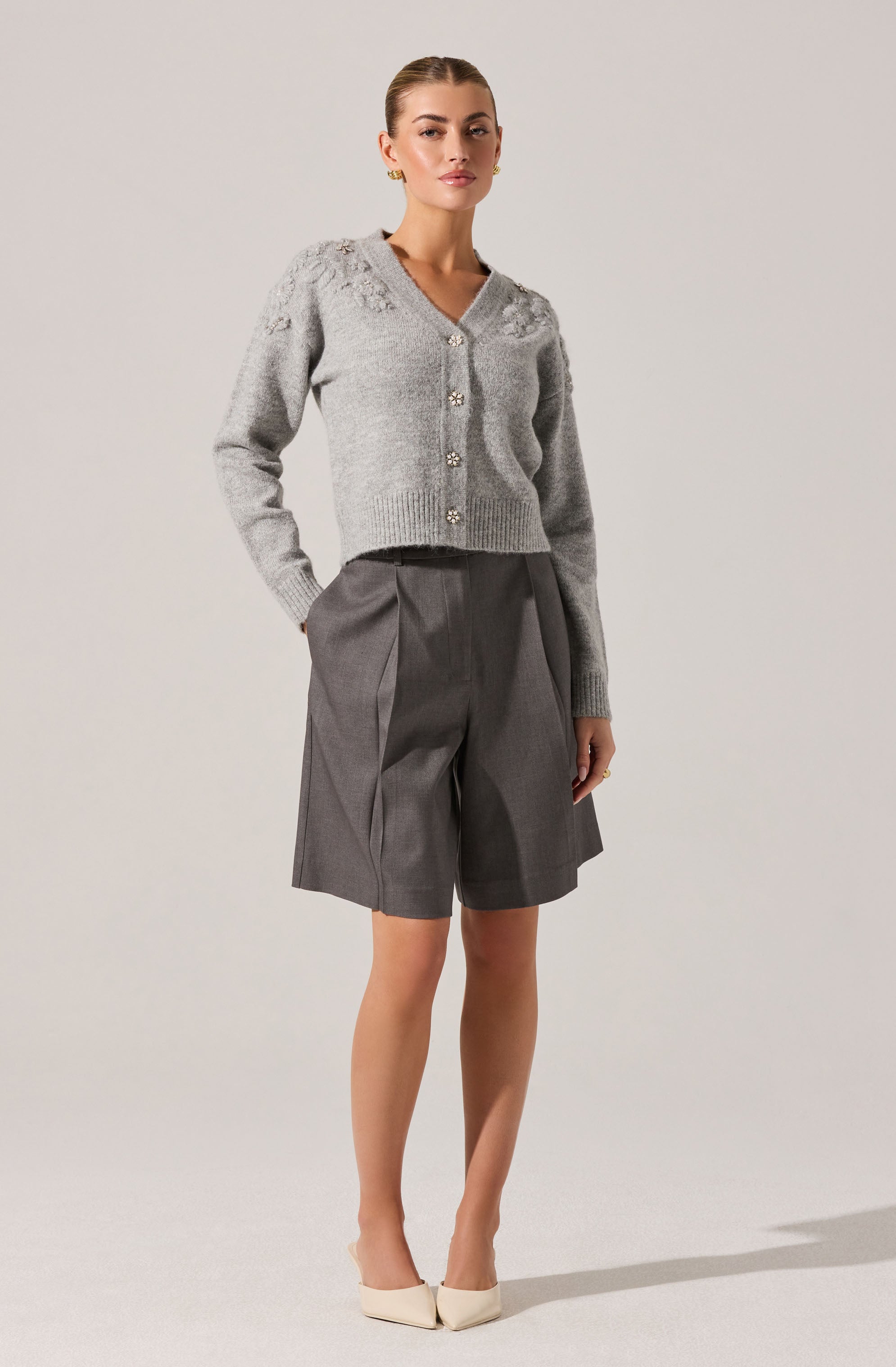 A woman is wearing a light gray, long-sleeved, v-neck cardigan with decorative buttons, paired with high-waisted, dark gray pleated shorts that have a loose fit. She is also wearing beige pointed-toe heels.