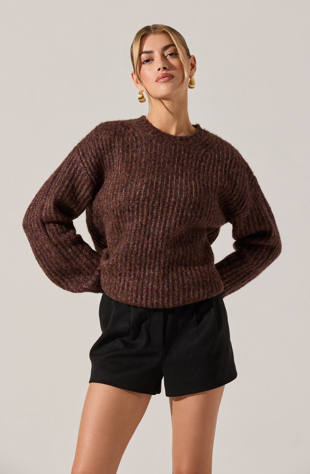 A woman is wearing a chunky, long-sleeved, brown knitted sweater with a textured pattern, and black high-waisted shorts with a loose fit. She has gold hoop earrings and styled blonde hair.