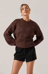 A woman is wearing a chunky, long-sleeved, brown knitted sweater with a textured pattern, and black high-waisted shorts with a loose fit. She has gold hoop earrings and styled blonde hair.
