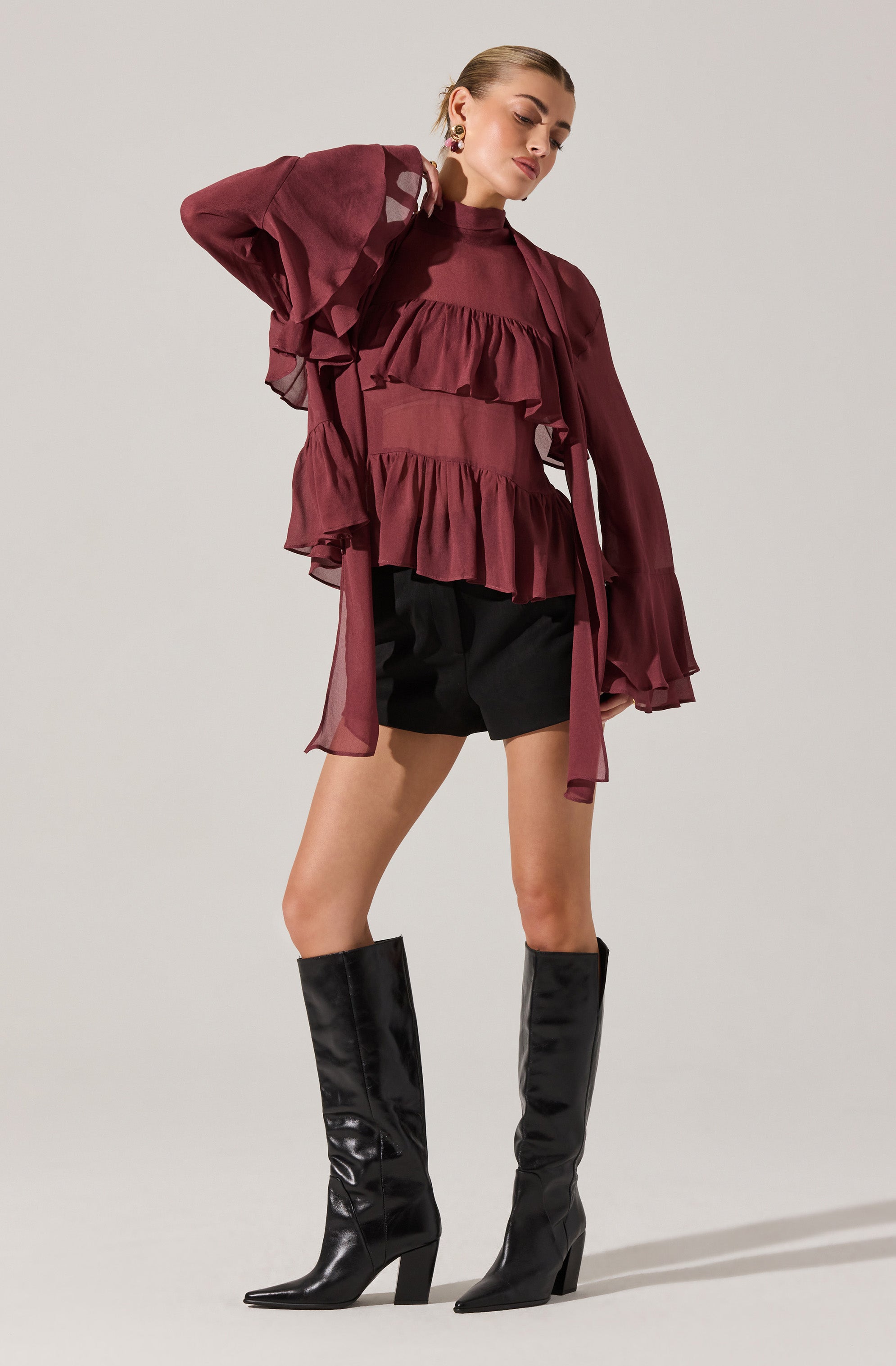 A woman is wearing a long-sleeved, high-neck, burgundy blouse with ruffled layers across the chest and front. The sleeves are sheer and billowy, gathered at the cuffs. She pairs the blouse with black shorts and tall, shiny black leather boots with high heels.