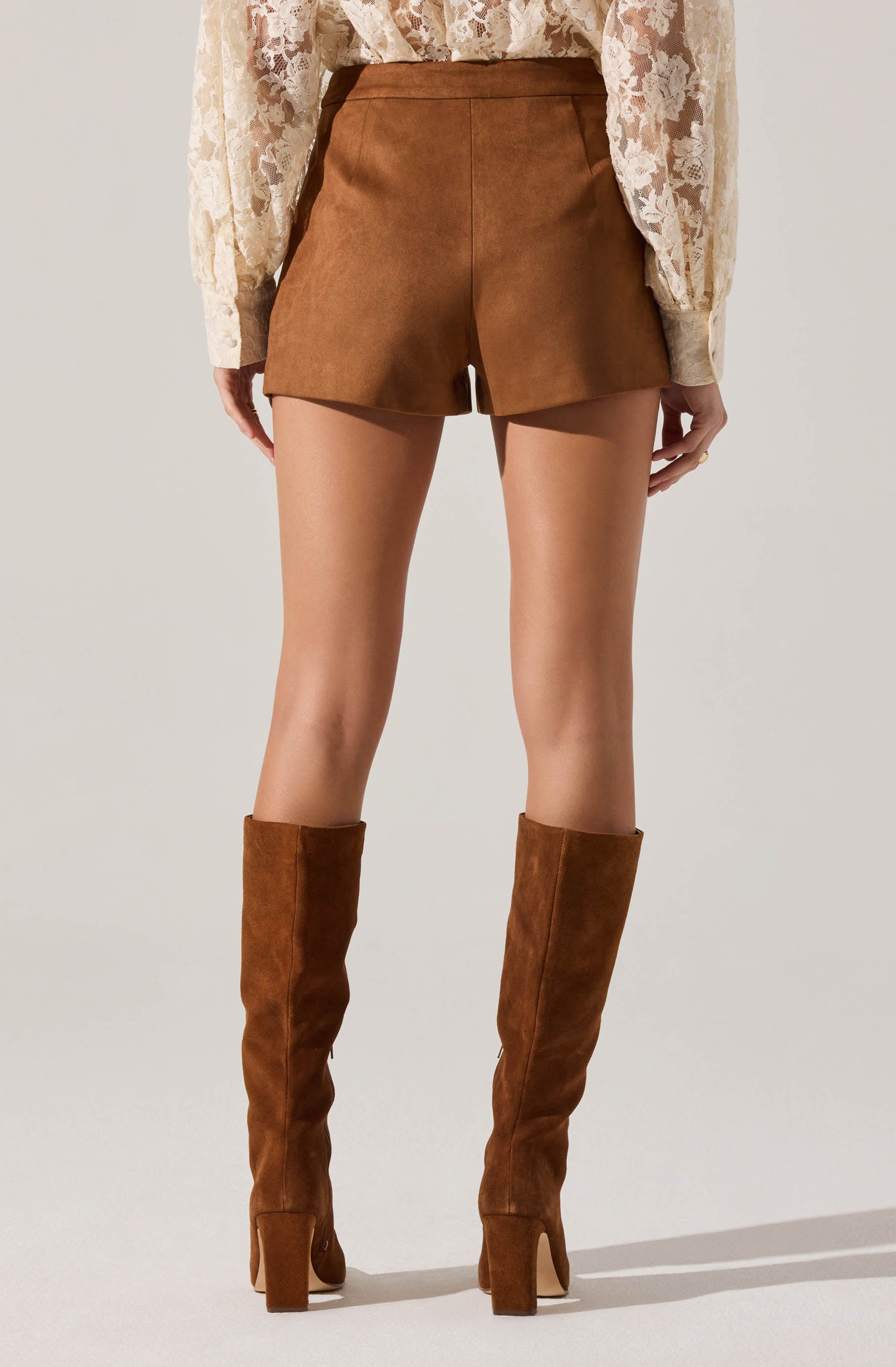 A person is wearing high-waisted brown shorts made of a suede-like material paired with tall, matching brown suede boots with block heels. The outfit is complemented by a long-sleeved blouse featuring a delicate cream-colored lace pattern.