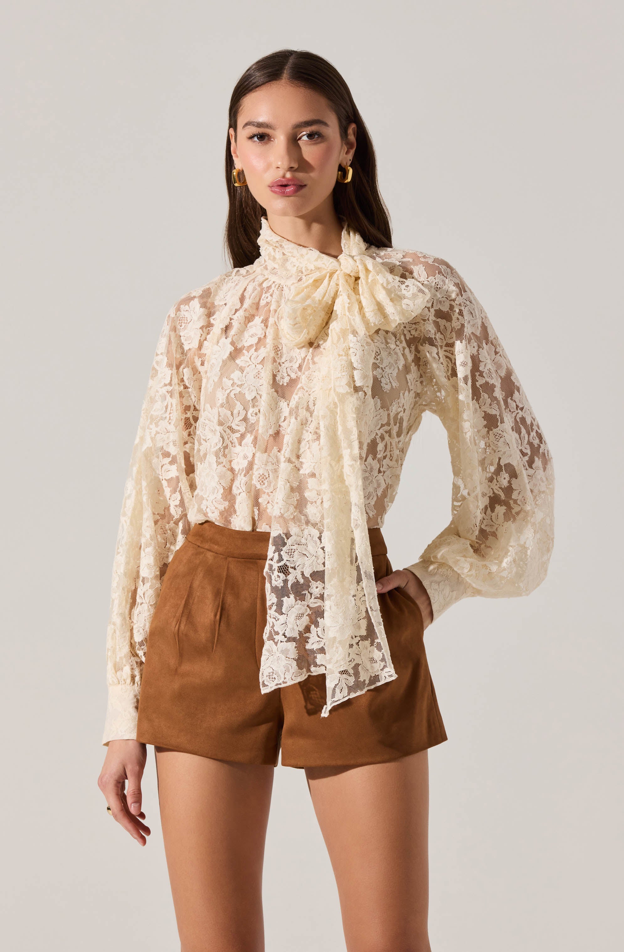 A woman is wearing a long-sleeved, sheer, cream-colored lace blouse with a large bow at the neckline. The blouse has a floral lace pattern and billowy sleeves that are gathered at the cuffs. She pairs it with high-waisted, brown suede shorts. She is accessorized with gold hoop earrings.