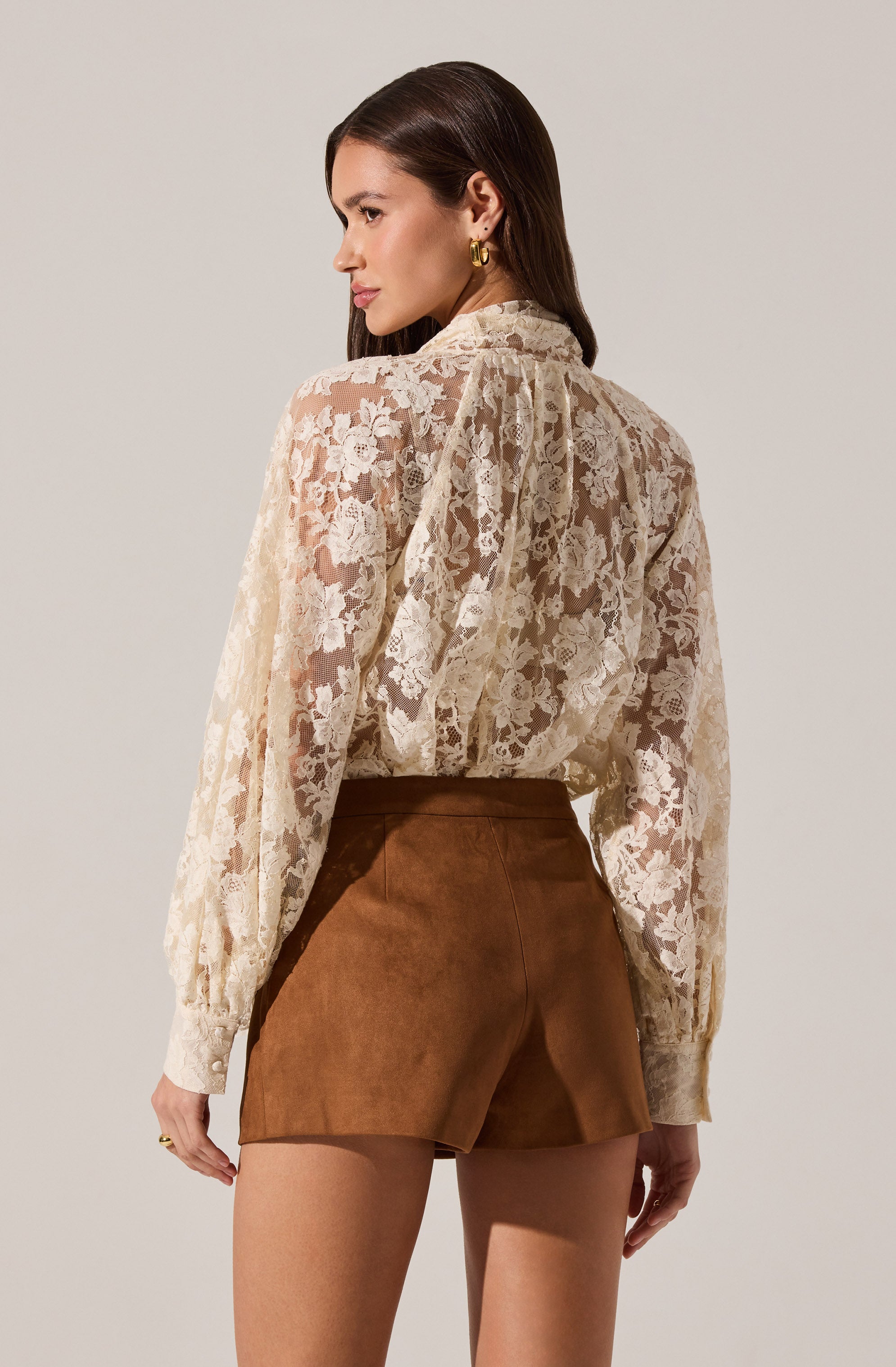 A woman is wearing a long-sleeved, cream-colored lace blouse with floral patterns. The blouse has a high neckline with a collar and puffed sleeves that gather at the wrists with buttoned cuffs. She pairs it with high-waisted brown shorts that have a smooth, suede-like texture. She is accessorized with gold hoop earrings.