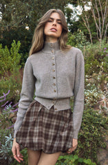 A young woman is wearing a light gray, long-sleeved cardigan with a high collar and button closure down the front. She pairs it with a high-waisted, brown plaid skirt with pleats, which features a subtle tartan pattern. The background is a lush outdoor setting with various green plants, trees, and flowers.