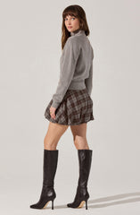 The woman is wearing a light grey, long-sleeved, cropped turtleneck sweater with a relaxed fit. She has on a short, pleated skirt with a brown and grey plaid pattern. She is also wearing tall, black, knee-high leather boots with pointed toes and stiletto heels.