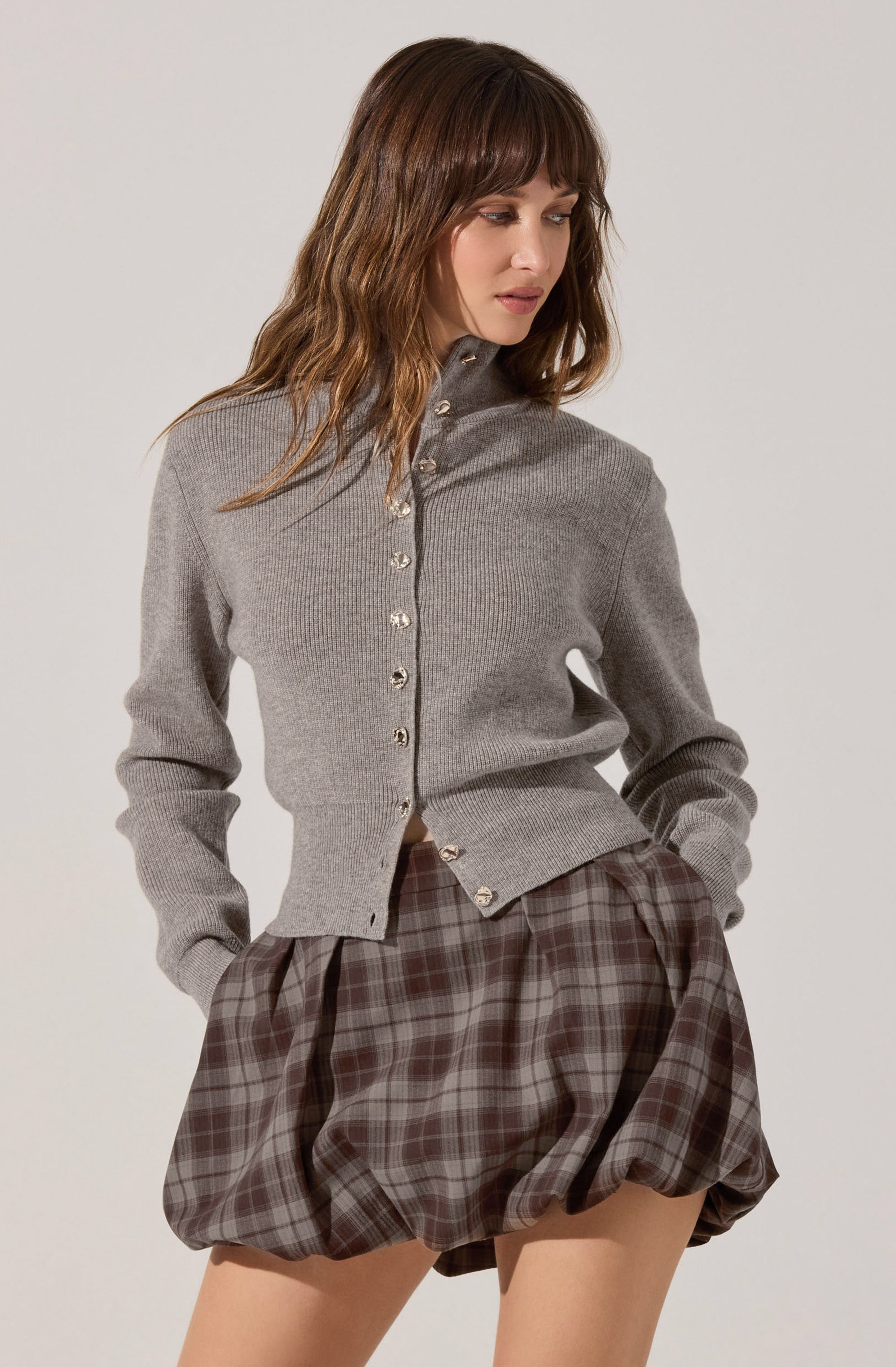 A woman is wearing a light gray, long-sleeved, ribbed knit cardigan with a high collar and pearl buttons down the front. She is also wearing a pair of loose-fitting, brown plaid shorts with a gathered, puffy style.