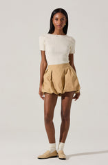 A woman is wearing a fitted, short-sleeved, ribbed, cream-colored top with slightly puffed sleeves. She pairs it with high-waisted, beige, pleated shorts that are loose-fitting and have a unique, rounded, and gathered design. She completes her outfit with light-colored socks and beige slip-on shoes.