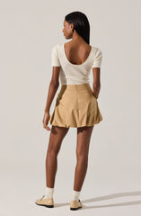 A woman is wearing a fitted, cream-colored, ribbed knit top with a low, rounded back and short sleeves. She pairs it with a beige, high-waisted, pleated mini skirt with a zipper at the back. She completes her outfit with cream-colored ankle socks and matching beige shoes. She has gold earrings and has long, straight black hair.