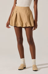 A person is wearing a high-waisted beige pleated skirt with large, gathered pleats creating a balloon effect. They are also wearing a sleeveless, ribbed, beige top tucked into the skirt. The outfit is complemented with beige loafers and cream-colored socks, and the person has a ring on their right hand.