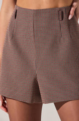 A person is wearing high-waisted shorts with a small checkered pattern in red, black, and beige. The shorts have belt loops at the waistband and side seam pockets. The fabric appears to be lightweight, suitable for warm weather. The person's hand is resting in the right pocket.