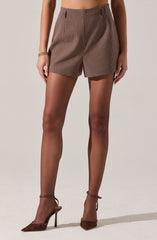 A person is wearing high-waisted shorts with a small checkered pattern in shades of brown, black, and white. The shorts have belt loops but no belt is worn. They are paired with sheer, nude-colored hosiery and brown pointed-toe high heels with ankle straps. The person's left hand is resting by their side, adorned with a gold bracelet.
