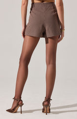 The image shows a person wearing high-waisted shorts with a small plaid check pattern in shades of brown and black. They are pairing the shorts with sheer nude stockings and brown high-heeled sandals that have ankle straps. One hand is resting by their side, wearing a gold watch and ring.