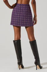 A woman is wearing a high-waisted, above-the-knee skirt made of a textured, woven tweed fabric with a purple, pink, and white checkered pattern. She is also wearing black, knee-high leather boots with stiletto heels.
