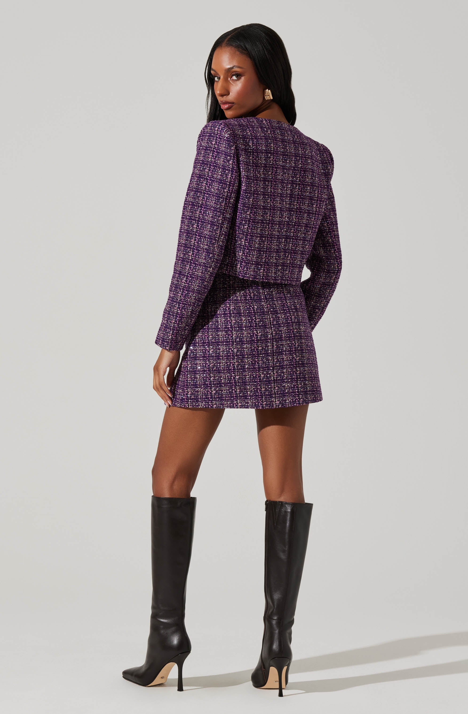 A woman is wearing a matching purple and black tweed blazer and mini skirt set. The blazer has long sleeves and a textured pattern, and the skirt is fitted and short. She is also wearing tall black leather knee-high boots with stiletto heels.