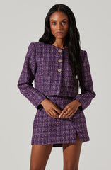 A woman is wearing a matching two-piece outfit made of purple and black tweed fabric. The top is a cropped, long-sleeved jacket with a rounded neckline and four decorative gold buttons down the front. The matching skirt is a short, fitted mini skirt with a small slit on one side.