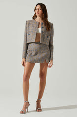 A woman is wearing a matching suit consisting of a cropped, long-sleeve blazer with a textured, multicolored fabric, and a mini skirt with a similar pattern and a slight slit on the front. She has on a white crop top underneath the blazer. She is also wearing high-heeled sandals with a bow detail.