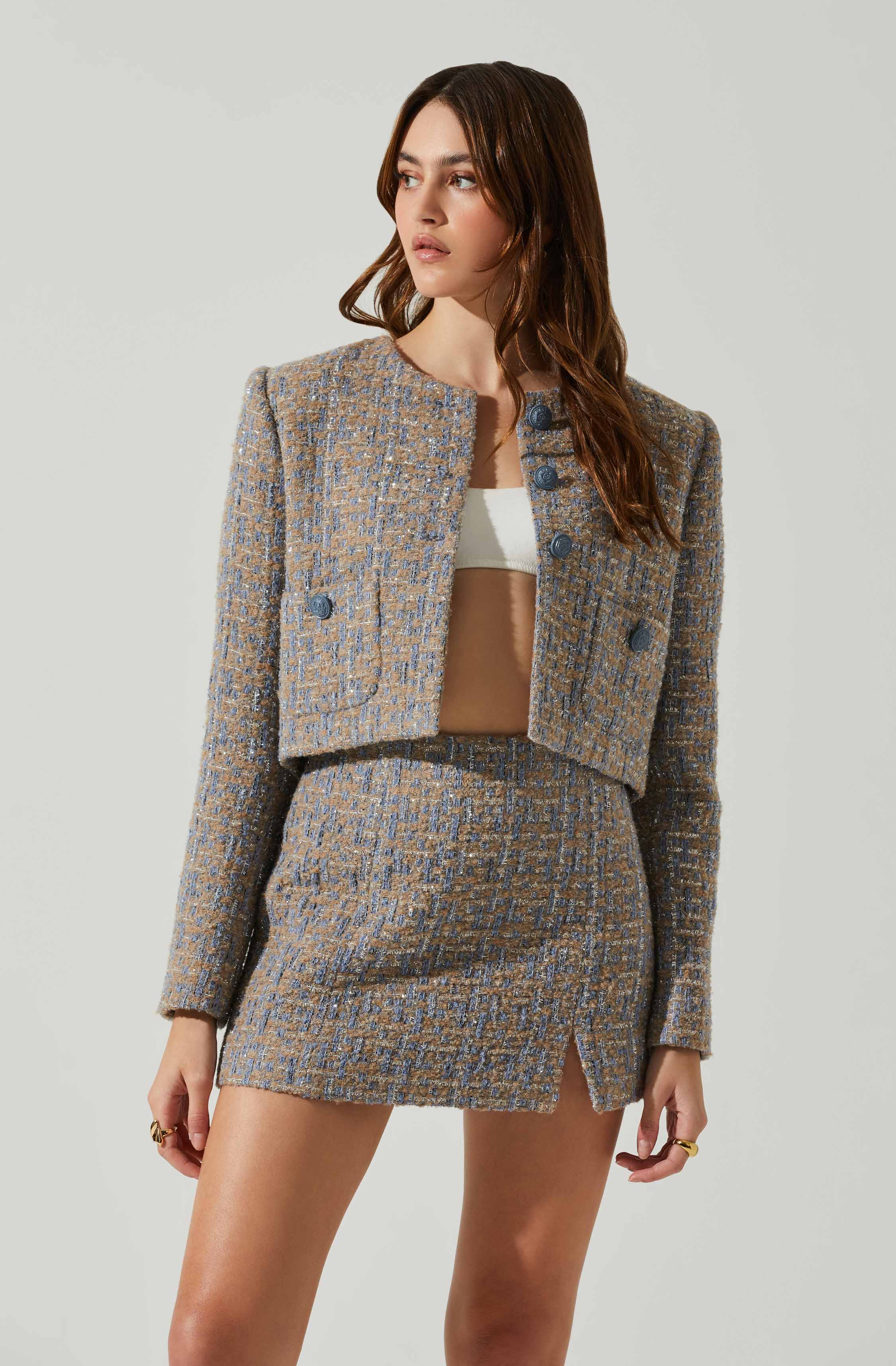 A woman is wearing a coordinated outfit consisting of a cropped, double-breasted blazer and a matching mini skirt, both made of a textured, patterned fabric in shades of brown and blue. The blazer has structured shoulders and large buttons, while the mini skirt features a small slit at the front. She is also wearing a white crop top underneath the blazer.