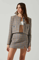 A woman is wearing a coordinated outfit consisting of a cropped, double-breasted blazer and a matching mini skirt, both made of a textured, patterned fabric in shades of brown and blue. The blazer has structured shoulders and large buttons, while the mini skirt features a small slit at the front. She is also wearing a white crop top underneath the blazer.