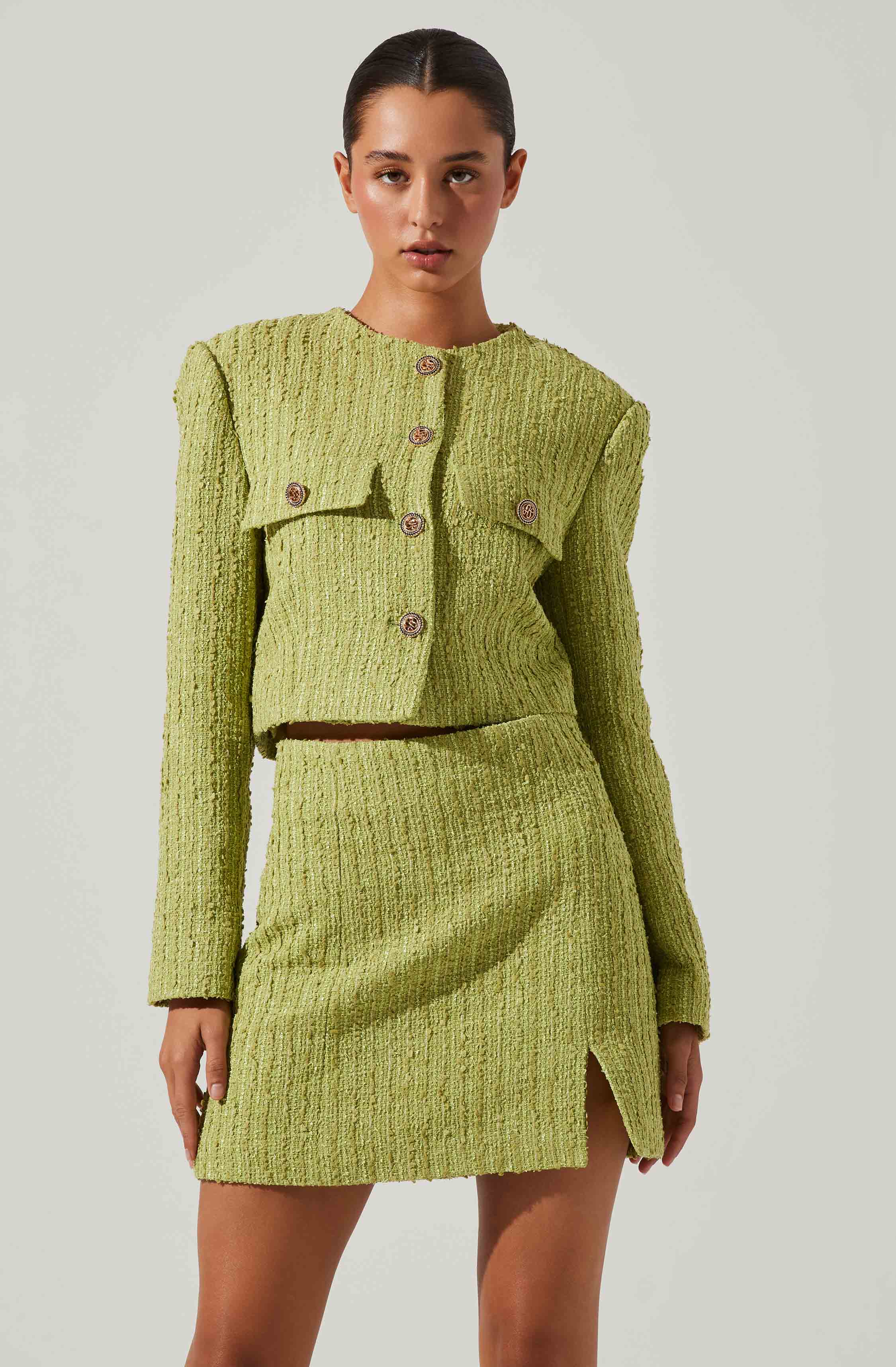 A woman is wearing a matching lime green textured two-piece outfit consisting of a cropped jacket and a high-waisted skirt. The jacket features a rounded neckline, long sleeves, and large decorative buttons down the front. The skirt has a slight slit on the left side. The fabric has a woven appearance with a subtle pattern.