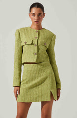 A woman is wearing a matching lime green textured two-piece outfit consisting of a cropped jacket and a high-waisted skirt. The jacket features a rounded neckline, long sleeves, and large decorative buttons down the front. The skirt has a slight slit on the left side. The fabric has a woven appearance with a subtle pattern.