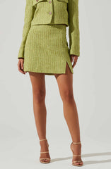 A person is wearing a matching light green textured skirt and jacket set. The skirt has a slit on the side and falls above the knees. They are also wearing beige high-heeled sandals with ankle straps.