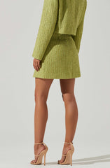 A person is wearing a matching two-piece outfit in a textured, lime green fabric. The outfit includes a long-sleeved top and a skirt that falls above the knees. The individual is also wearing nude-colored high-heeled sandals with ankle straps.