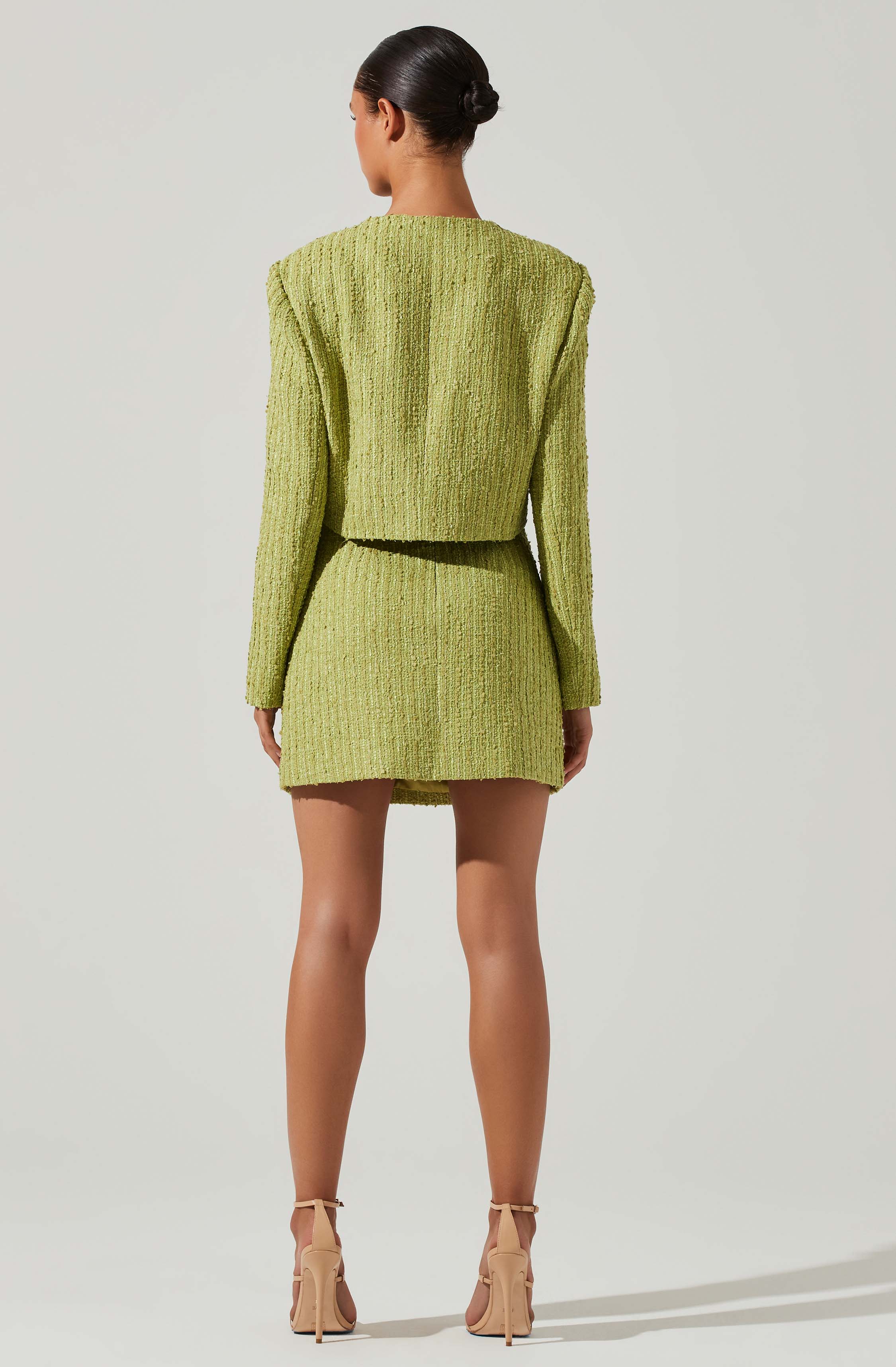 A woman is wearing a matching light green textured jacket and skirt set made of a woven or tweed-like fabric. The jacket is cropped and loose-fitting, ending above the waist, while the skirt is high-waisted and short. She is also wearing nude high-heeled sandals with ankle straps. Her hair is styled in a neat bun.