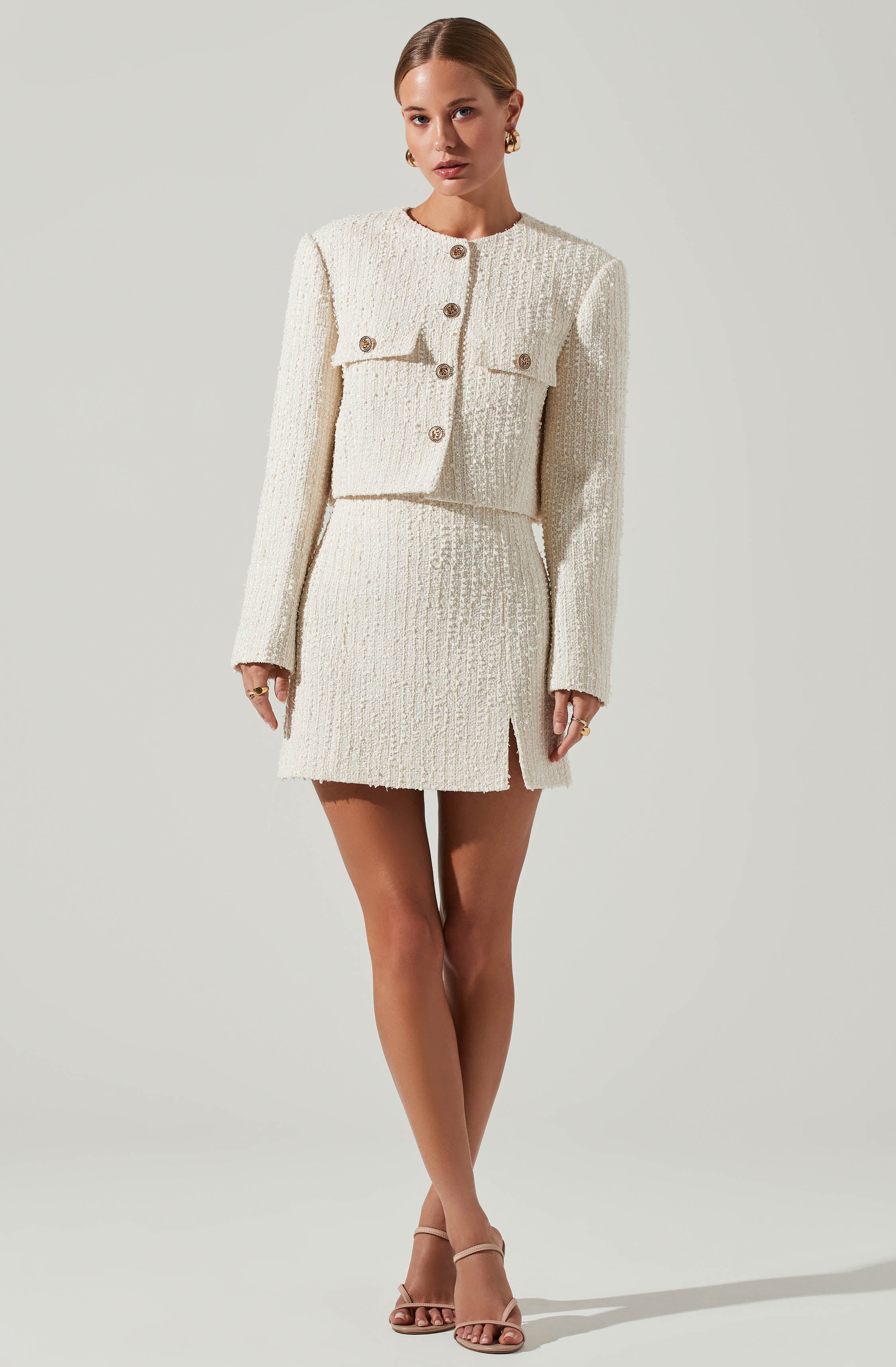 A woman is wearing a cream-colored, textured matching set consisting of a cropped jacket and a high-waisted skirt. The jacket has long sleeves, a rounded neckline, and decorative buttons down the front. The skirt is fitted, with a small slit on one side. The fabric appears to be a boucle or tweed material, giving it a classic, elegant look.