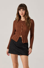 A woman is wearing a long-sleeved, brown, cable-knit cardigan with a rounded lace collar and white buttons down the front. She pairs it with a black, high-waisted mini skirt.