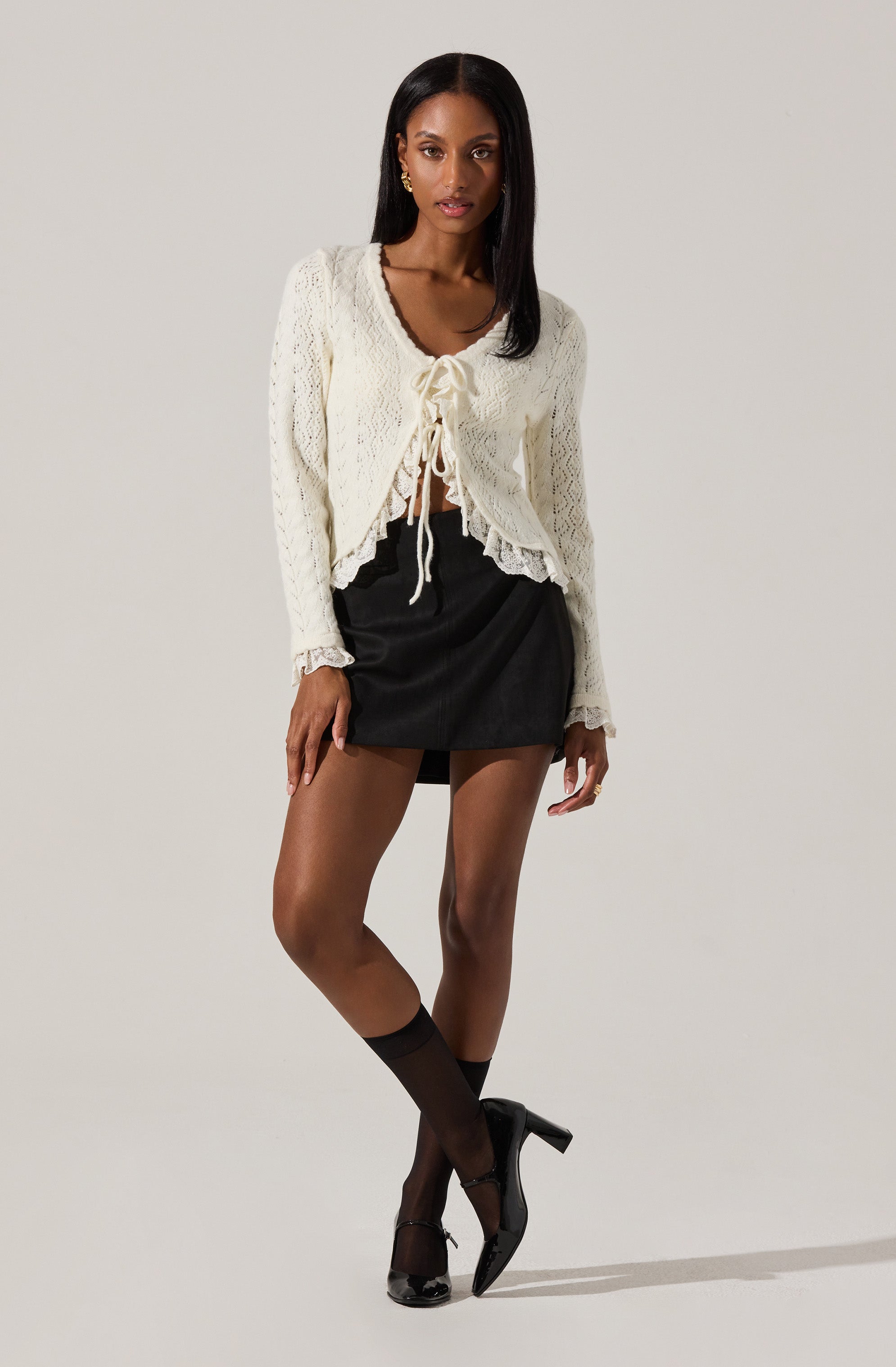 A woman is wearing a cream-colored, long-sleeved, knitted cardigan with a ruffled hem and a bow tie at the front. She pairs it with a black mini skirt, sheer black ankle socks, and black high-heeled shoes.