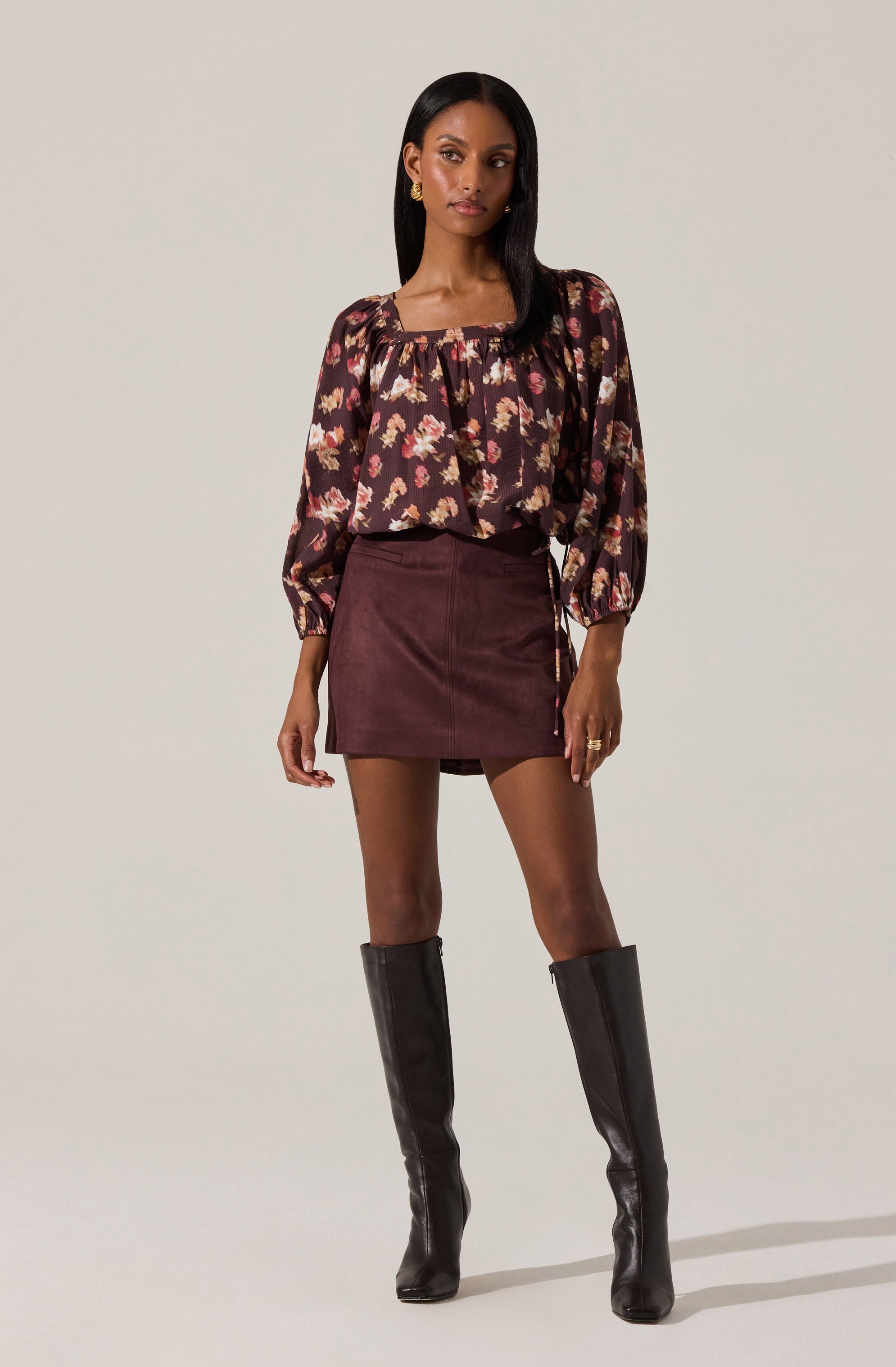 A woman is wearing a long-sleeved blouse with a floral pattern in shades of pink, beige, and white on a dark background. The blouse has a square neckline with a slight ruffle detail and puffed sleeves gathered at the wrists. She pairs it with a fitted, high-waisted maroon skirt that falls mid-thigh. She is also wearing tall black leather knee-high boots. She has gold hoop earrings and a ring on her right hand.