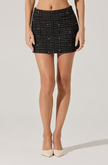 A woman is wearing a black, textured mini skirt with a grid pattern of white and beige lines, accented with small metallic beads or embellishments. She is also wearing nude-colored high heels.