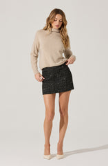 A woman is wearing a beige, high-neck, long-sleeve sweater made of soft, cozy material. She is also wearing a black mini skirt with a checkered pattern featuring white and red accents. The outfit is paired with off-white pointed-toe heels.