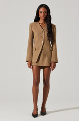 A woman is wearing a tan-colored, tailored blazer with shoulder pads and four buttons down the front. She has on a matching tan mini skirt, finished with black pointed-toe heels.