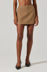 A person is wearing a high-waisted, camel-colored wool mini skirt with front slit and two small horizontal welt pockets at the front. They are also wearing black pointed-toe stiletto heels with a bow detail on the front.