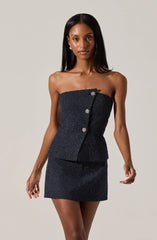 A woman is wearing a strapless, dark-colored, sparkly mini dress with a subtle shimmer. The dress features a vertical row of decorative buttons along the front, adding an elegant touch. The fabric appears textured with a glittering effect, giving the outfit a sophisticated and glamorous look.