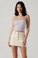 A woman is wearing a light purple, knitted, sleeveless crop top with lace-trimmed straps and a pattern of vertical and horizontal lines. She pairs the top with a high-waisted, cream-colored leather mini skirt that has belt loops and a front seam.