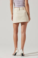 A woman is wearing a light purple sleeveless knit top with a subtle diamond pattern, a high-waisted cream-colored mini skirt with belt loops, and white high-heeled sandals with ankle straps.