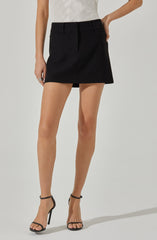 A person is wearing a high-waisted, black A-line mini skirt with belt loops, paired with open-toe, black strappy high-heeled sandals. They are also wearing a short-sleeved, cream-colored top with a subtle textured pattern.