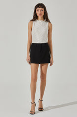 A woman is wearing a sleeveless, light-colored top with a subtle vertical stripe pattern and ruffled neckline paired with a high-waisted black mini skirt. She has on black high-heeled sandals with ankle straps.