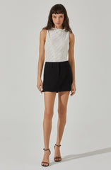 A woman is wearing a sleeveless, white textured top with a high neckline and slightly ruffled edges. She has paired the top with a high-waisted, black A-line mini skirt that features belt loops and a subtle seam detail at the front. She is also wearing black strappy high-heeled sandals.