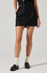 A person is wearing a black sleeveless button-up top and a high-waisted black skirt that falls above the knees. They have on beige socks and black loafers with a shiny finish.