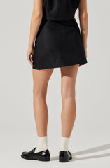 A woman is wearing a sleeveless black top tucked into a high-waisted black denim skirt with visible back pockets. She has on cream-colored socks and shiny black loafers.