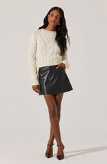 A woman is wearing a cream-colored, chunky knit sweater with cable patterns, paired with a high-waisted black leather A-line mini skirt. She also has on dark brown high-heeled shoes with ankle straps.