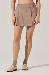 A person is wearing beige high-waisted shorts with pleats and a side tie detail. They are also wearing beige shoes with a rounded toe and flat heel, and beige socks that are slightly scrunched down.